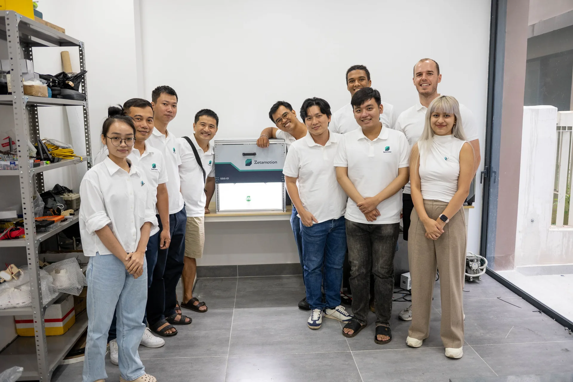Group of diverse team members in white shirts posing in a workshop around a Zetamotion device on a table in front of them.