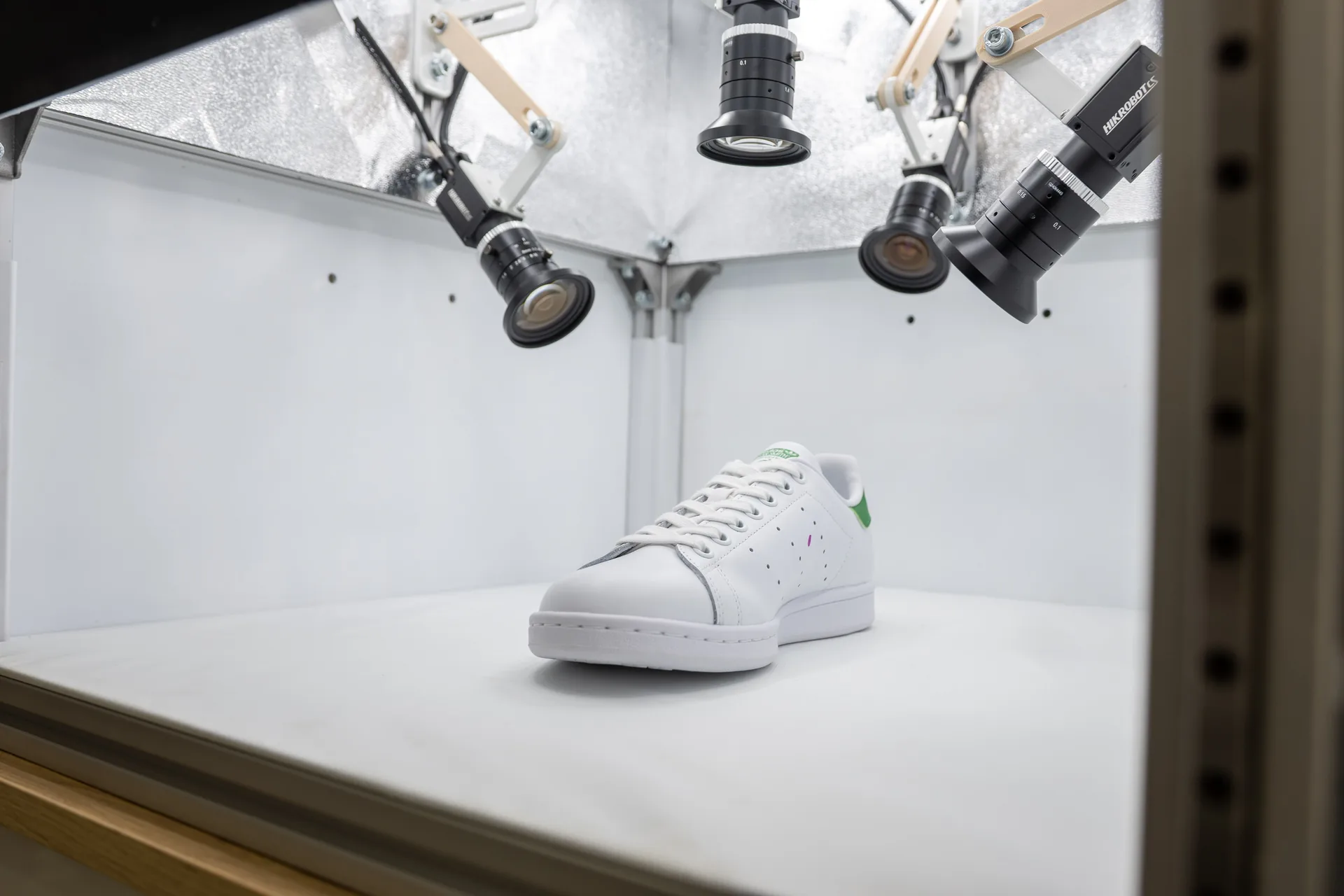 White sneaker on a white platform beneath robotic arms in a high-tech lab/production setup, suggesting automated inspection or handling.
