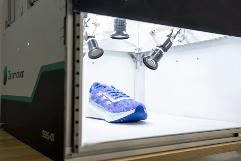Blue running shoe placed inside a motion-testing chamber, with robotic arms/cameras above for analysis in a lab.