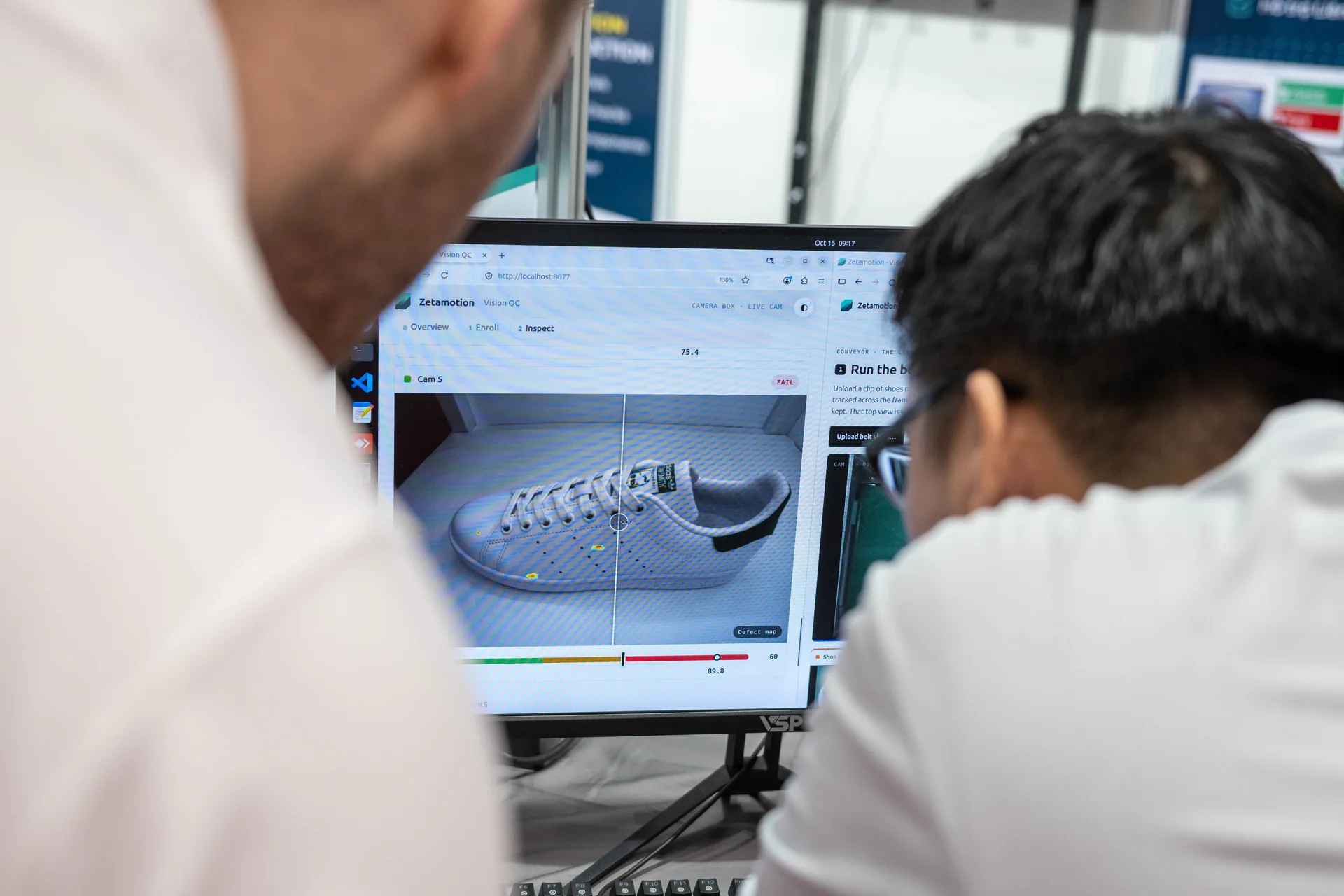 Two people view a computer monitor displaying a 3D shoe design with editing tools on screen, in a busy work environment.
